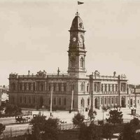 Image: a large two storey stone public building with a tall clock tower protruding from one corner stands on the corner of two city streets. A large park can be seen in the foreground of the image. 