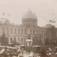 Image: a large crowd of people in 1880s clothing gather in front of a large building with a domed roof watching as horse drawn carriages pass by.
