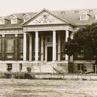 Image: A large rectangular brick building with Greek Revival-style columns flanking its main entrance