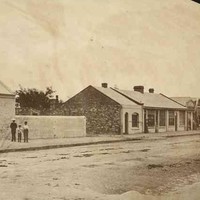 Image: men and boys in 1860s attire stand on a paved sidewalk outside their cottages. 
