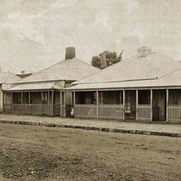 Image: a row of three identical brick, iron-roofed cottages with fenced in verandahs. A group of four children stand with their mother outside the rightmost cottage. On the far left is a fourth building which is being used as a general store.