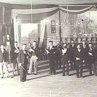 Image: a scattering of men in a large room dressed with lines of chairs and featuring a small stage with a piano.