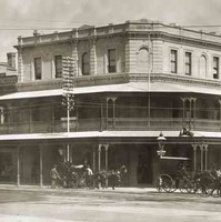 Image: a three storey corner hotel with a protruding balcony on the second floor which also forms a verandah. Horse drawn vehicles pass on the street outside. 
