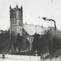 Image: A large stone church building with a tower topped by four spires stands at the corner of two dirt roads. A man stands in front of the churchyard fence
