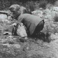 Image: A man in a hat and outdoor clothing is poised on all-fours and looking intently at the ground. A light-coloured bag is on the ground next to him