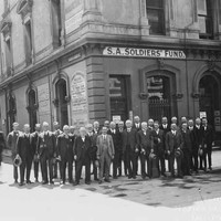 Image: A group of men wearing 1920s era suits and holding their hats pose for a photograph on a street corner outside a building with a sign reading: "S.A. Soldiers' Fund". To one side are two more men who only have one leg. 