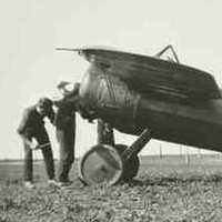 Image: Three men stand next to a First World War-era monoplane parked on a large, flat expanse of ground