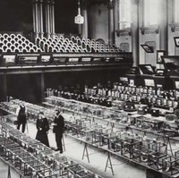 Image: Two men and a woman stand in a large open room with four large tables arrayed in rows. On each table are several cages, each of which contains one live bird