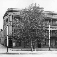 Image: a large tree obscures the view of a three storey stone building on the corner of a wide street and small alley. The building has balconies on its second and third floor and a corner door at street level. 