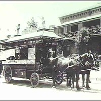 Image: a man in early 20th century clothing stands by a horse drawn tram which is parked in front of a two storey house.