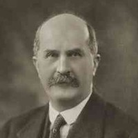 Image: A photographic portrait of a balding, moustachioed middle-aged man wearing a suit and tie