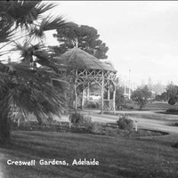 Image: Black and white photograph of a rustic wooden rotunda situated in public gardens. Indistinct figures wander along the garden paths in the middle-ground while buildings can be seen in the distance.