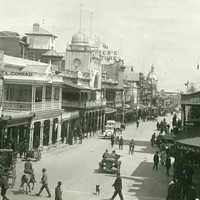 Image: A view of a city street with many people in 1920s attire walking, riding horses or driving motor vehicles. The street is lined with 2 and 3 storey commercial buildings, most with verandahs and/or balconies and a number featuring towers with domes. 
