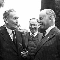 Image: Three men in suits and ties speak with one another on a property's well-manicured grounds. Each man is smiling, suggested he is amused by the conversation