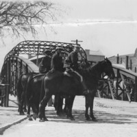 Image: Several men on horseback form two ranks in front of an iron bridge