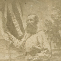 Image: Photograph of bearded man standing in front of a painted background of a beach. He stands in three quarter profile, legs crossed, and holds a flag in his right hand. His hat sits on the ground in front of him.