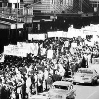Image: a large group of people holding protest signs against the Vietnam War march down a city street