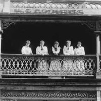 Image: a group of five women in white dresses in an early 20th century style stand on a balcony of a two storey building which has signs declaring it the Globe Dining Rooms