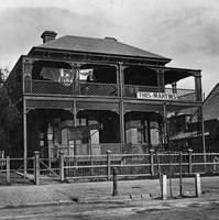 Image: a two storey building with a combination verandah and balcony, decorated with lattice work, sits behind a wooden fence. Washing dries on a line on the balcony.