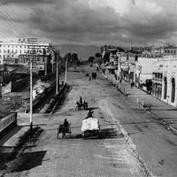 Image: dark clouds hang over a wide dirt city street which is lined with a variety of buildings from single storey tin sheds to large stone constructions of up to six storeys. Travelling down the road are a range of horse drawn vehicles. 