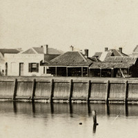 Image: A line of buildings sit atop a long wharf. In front of the wharf is a river, in which three small boats are moored