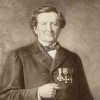 Image: An old photograph of a man wearing a dark coat, bow-tie and medals posing with one hand holding a book upright on a table by his side, the other, his right, tucked in between the buttons of his coat