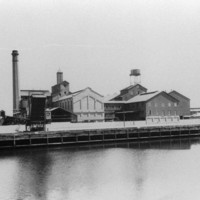 Image: A complex of multi-storey brick buildings and large, corrugated metal-clad warehouses fronted by a river. A large brick chimney is visible at one end of the complex