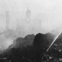 Image: A brick chimney and a few brick multi-storey buildings are shrouded in smoke from a fire. The burning sugar stock is visible in the foreground