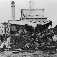 Image: Destroyed and partially-destroyed buildings are visible behind a large, burnt pile gunny sacks interspersed with twisted sheets of corrugated metal