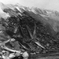 Image: A large, smouldering pile of charred gunny sacks, atop which are fragments of burned corrugated metal sheeting