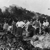 Image: A group of men dressed in work clothes and wide-brimmed hats pose for a photograph next to piles of fire-damaged sacks