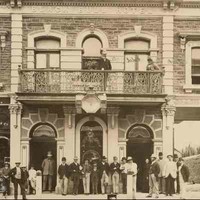 Image: A group of at least 24 men in 1880s attire stand on the street outside a two storey stone terrace building. A further two figures can be seen on the balcony above. The building features decorative window surrounds and extends over a large archway.