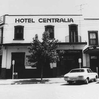 Image: a two storey corner hotel, painted white with dark windows and doors and a flat roof. Small, round balconetes protrude from some of the upper floor openings. 1980s era cars are parked on the street outside.