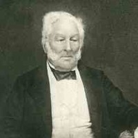 Image: a photograph of a seated, white haired man with a beard. His left hand is resting on a table and his right is on his lap. He is wearing grey trousers, a white shirt and waistcoat and a black jacket and bow tie. 