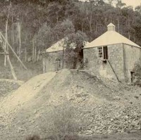 Image: Two single-storey stone buildings stand next to large dirt piles and a wooden tower