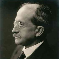 Image: A photographic portrait of a moustachioed man in a suit and wire-rimmed spectacles. He is shown in partial profile