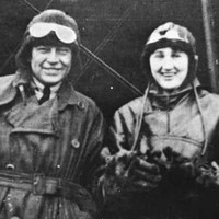 Image: A man and woman wearing First World War-era pilot’s clothing and headgear stand in front of a biplane
