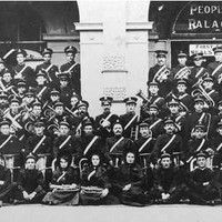Image: Members of a large band, dressed in dark uniforms with white sashes, pose in lines with their instruments outside of a building with arched doorways.