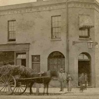 Image: A man, woman and children in 1870s dress stand on the street beside a horse drawn cart laden with straw, outside of a two storey hotel with a corner door, flat roof, arched ground floor windows and awnings over the rectangular second storey windows