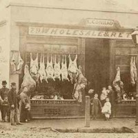 Image: a group of men, women and children in 1860s attire stand outside a single storey building which is operating as a butchers. A range of whole animal carcases are hung outside the store while other cuts of meat can be seen in a window display. 