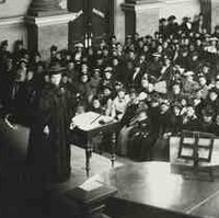 Image: A woman in cap and gown stands on a stage in front of a large audience of women dressed in late Victorian-era attire