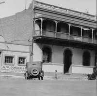 Image: A 1930s-era automobile is parked on a street in front of two buildings. One building is two storeys, made of brick, and has a second-floor verandah