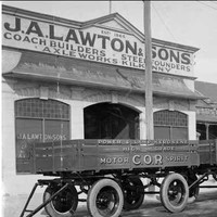 Image: three four wheeled trailers are parked in a row outside of a single storey building with arched windows and open entranceway. A parapet sign on the building reads: "J.A. Lawton & Sons. Coach builders, Steelfounders, Axelworks, Kilkenny"