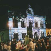 Image: a crowd gathers in front of a gothic style building which is lit up by a projection and glows purple and green