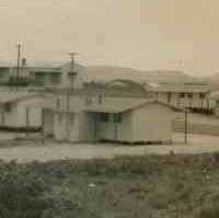 Image: A group of single-storey prefabricated buildings are situated on a remote piece of scrubby land. Low hills are visible in the distant background