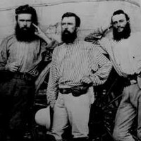Image: four bearded men stand in front of a covered wagon