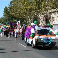 Image: women carrying banners and holding green and purple balloons march down a city street following a van decked out in balloons, bows and swathes of coloured fabric. One of the women near the front is beating a large drum
