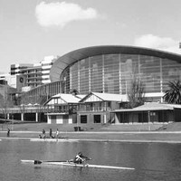Image: rowers travel down a wide river with grassy banks in front of a 24 storey skyscraper and a large convention centre with a multi-storey curved glass wall.