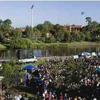 Image: a large group of people in modern dress stand in a park in front of a large screen. The park is also dotted with colourful marquees. To the right of the picture a rotunda is visible while in the background, across a lake, stadium lights can be seen