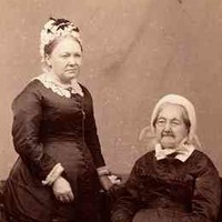 Image: sepia photograph of two women, one seated, one standing, wearing black dresses with white lace collars and white bonnets. The seated woman, the elder of the two, holds a book on her lap. 